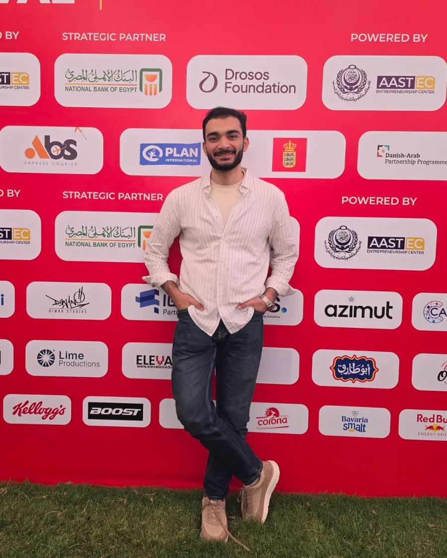 Ahmed Elagamy standing in front of the Rally Startup Festival sponsor wall at AAST